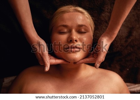 Partial image of female masseur doing face massage of woman in spa salon. Portrait of young blonde caucasian girl with closed eyes lying on massage table. Concept of rest with spa treatment. Top view