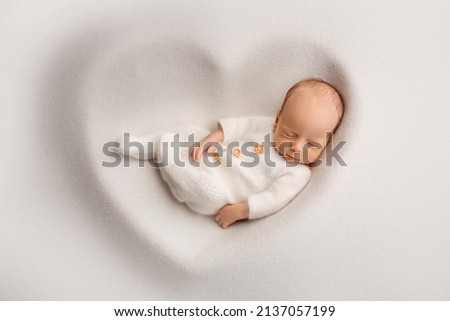 A sleeping newborn boy in the first days of life in a white soft cocoon with a knitted woolen white hat on a white background in the shape of a heart. Studio macro photography, portrait of a newborn. Royalty-Free Stock Photo #2137057199