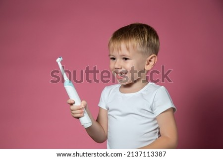 Smiling caucasian little boy holds electric sonic toothbrush on pink background.