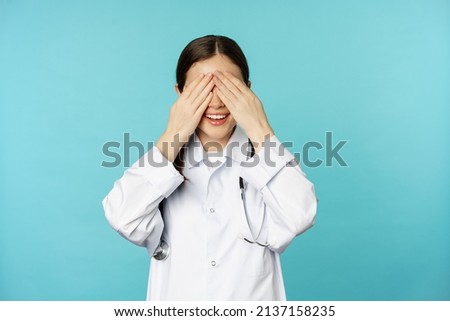 Smiling happy therapist, woman doctor cover close eyes with hands and waiting for surprised, anticipating, standing in white coat over torquoise background