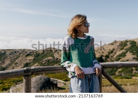 Outdoor portrait of European young woman wearing in stylish bright sweater in sunglasses is posing on the picturesque road against blue sky and green hills. High quality photo