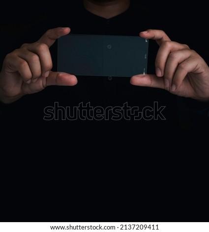 Future transparent glass phone Technology. Mockup blank horizontal screen futuristic super slim transparent smartphone in person's hands, taking photograph action, on black background with copy space.