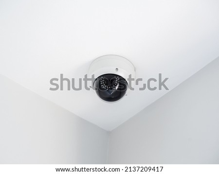 Digital security eye. A white round indoor CCTV surveillance camera monitoring security on ceiling in the room corner inside the white clean building.