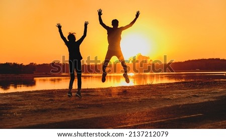 Male and female couple jumping happily at sunset with sun in the background, silhouette.
