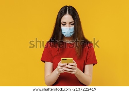 Cute young brunette woman 20s wears basic red t-shirt sterile face mask ppe to safe from coronavirus virus covid-19 flu hold in hand use mobile cell phone isolated yellow background studio portrait