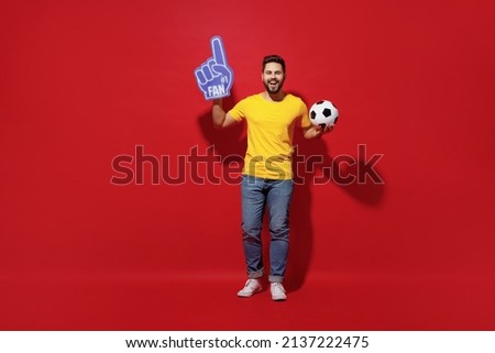 Full size body length young bearded man football fan in yellow t-shirt cheer up support favorite team hold soccer ball fan foam glove finger up isolated on plain dark red background studio portrait Royalty-Free Stock Photo #2137222475