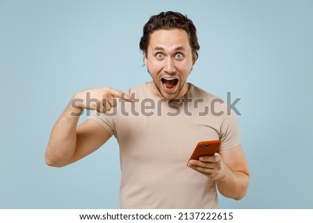 Young overjoyed excited caucasian man 20s in casual basic beige t-shirt point index finger on mobile cell phone chat online isolated on pastel blue background studio portrait. People lifestyle concept