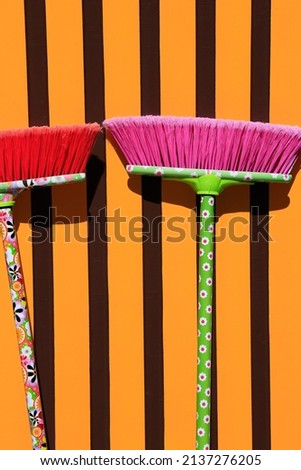Two broomsticks with red and pink bristles and colorful patterns in front of a wall with vertical geometric lines in orange and black. Spring cleaning, cleaning concept, empty space for text or logo
