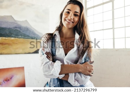 Cheerful artist smiling at the camera while standing in front of her paintings. Happy female painter standing with her arms crossed. Creative young woman displaying a collection of her artwork.