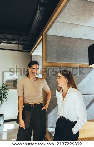 Successful businesswomen sharing a laugh in the office. Two young female entrepreneurs laughing cheerfully while standing together in a modern workspace. Royalty-Free Stock Photo #2137296809