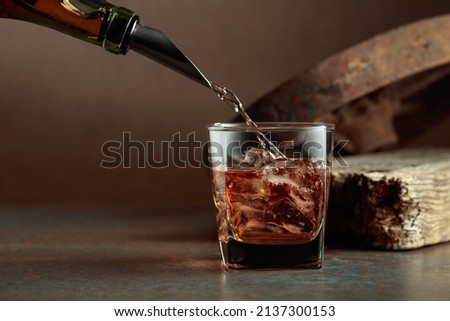 Whiskey is poured from a bottle into a frozen glass with natural ice. Rough rusty background.