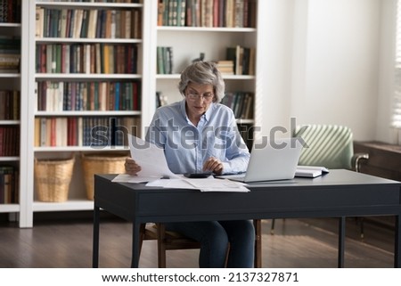 Mature accountant woman sit at workplace desk sorting out bills, making payments, calculates summary for pay through e-bank system on laptop. Checking and management of finances, accounting concept Royalty-Free Stock Photo #2137327871