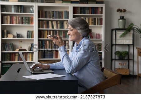 Older generation using modern tech, comfort communication, ai, workflow concept. Mature 60s business woman sit at workplace desk with laptop holds cellphone makes call talks to client on speakerphone