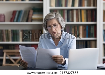 Serious thoughtful middle-aged woman in glasses looks worried read news in formal document sit at workplace desk with wireless computer. Older female review paper letter, learns report feels concerned Royalty-Free Stock Photo #2137327883