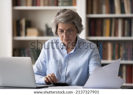 Middle-aged concentrated woman sits at table calculates summary for monthly payment, managing finances, work sit at workplace with laptop. Pay bills through e-bank system, money flow control concept Royalty-Free Stock Photo #2137327901