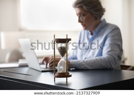 Busy productive mature businesswoman office employee work on laptop sit at workplace desk, hourglass on table, forefront view. Measure time, deadline, time management for business efficiency concept Royalty-Free Stock Photo #2137327909