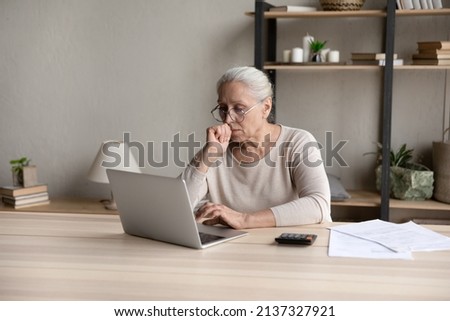 Aged woman sit at desk with calculator and papers, pay bills through e-bank application, looks at laptop screen feels concerned due bank debt or overdue. Older gen use tech, finances, troubles concept Royalty-Free Stock Photo #2137327921