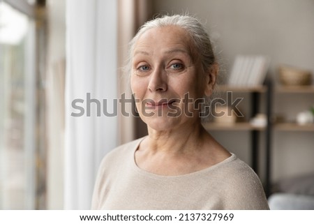 Head shot portrait attractive older woman standing in living room, staring at camera, looks satisfied feels carefree. Medical insurance cover for elderly citizen, untroubled life on retirement concept