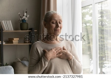 Smiling peaceful older female with folded palms on chest standing alone in living room with eyes closed praying or showing sign of gratitude. Believe, faith, charity and appreciation symbol concept Royalty-Free Stock Photo #2137327981