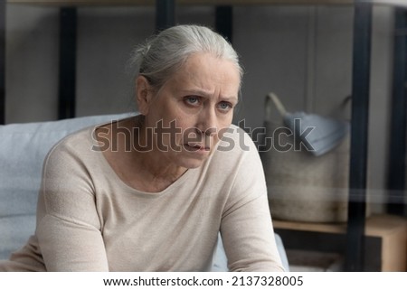 Pessimistic aged woman sits on sofa in living room feels hurt and lonely, lost in sad thoughts related to senile disease, mental disorder or dementia. Life troubles, solitude of older citizen concept Royalty-Free Stock Photo #2137328005