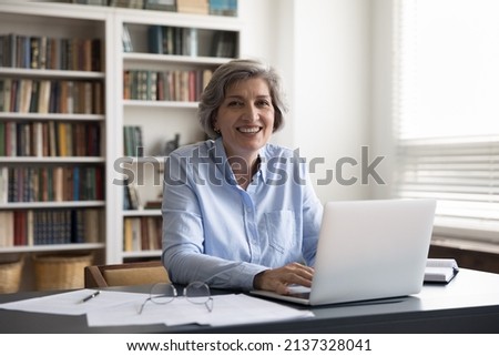 Portrait of smiling successful older 60s businesswoman, small business owner, sitting at workplace desk with computer staring at camera looking satisfied feels happy. Workday at modern office concept Royalty-Free Stock Photo #2137328041