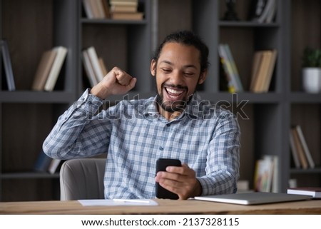 Excited happy Black guy using mobile phone at workplace table, feeling joy, getting good news, reading message on smartphone, celebrating win, success, achieve, making winner yes hand Royalty-Free Stock Photo #2137328115