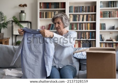 Dissatisfied client of retail services, complains, goods return concept. Aged 60s woman sit on sofa unpack received parcel box check female blouse looks disillusioned by fabric quality, obsolete model Royalty-Free Stock Photo #2137328159