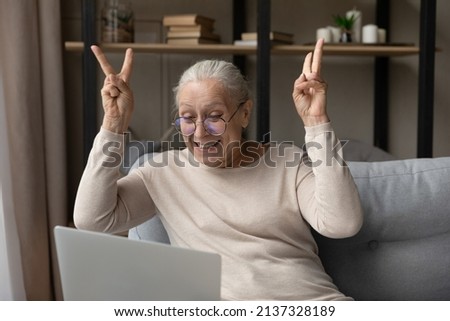 Senior woman in glasses sit on sofa staring at laptop, gesturing while make video call, greets grown up children looking overjoyed. Older generation use modern tech, remote communication, fun concept