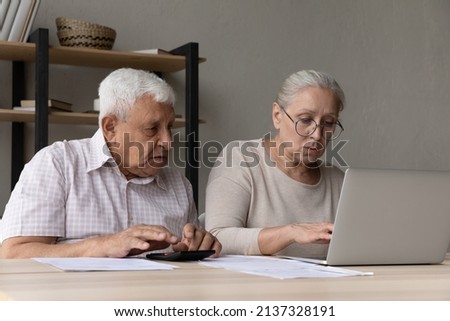 Serious aged spouses sit at desk use calculator make expenses summation, pay domestic bill use e-bank application on laptop, looking concerned due debt or huge taxes, manage household finances concept Royalty-Free Stock Photo #2137328191