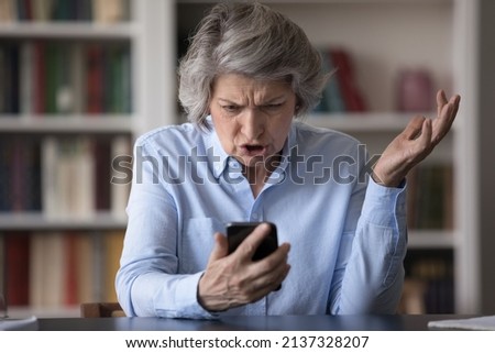 Angry older businesswoman sit at workplace staring at cellphone screen, feels mad looks shocked by awful news, dissatisfied by discharged or broken smartphone, get scam message, missed call concept Royalty-Free Stock Photo #2137328207