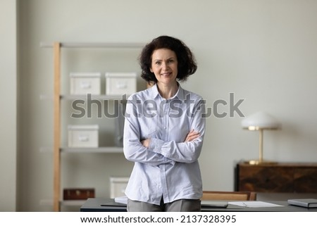 Confident mature business leader woman standing with hands folded at office workplace, looking at camera, smiling. Senior businesswoman, CEO, entrepreneur head shot portrait