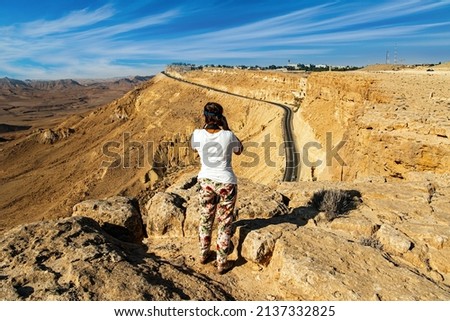 Woman photographing a beautiful landscape on a steep cliff. Israel. Crater Makhtesh Ramon in the Negev Desert declared a Geological Reserve. The morning after the starfall. Israel