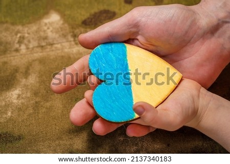 Man's and child's hand holding a heart painted in the colors of the flag of Ukraine, camouflage background