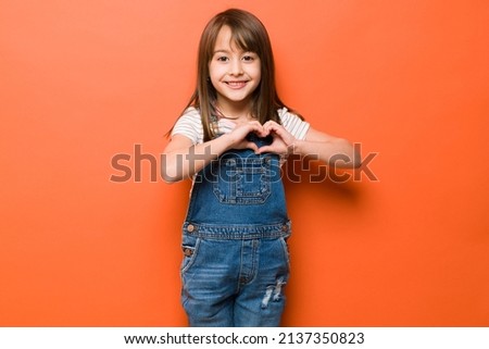 Adorable little girl making a heart shape with her hands and smiling in a studio