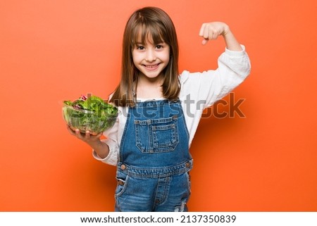Happy little Caucasian girl holding a bowl of salad and showing her strong arm for eating healthy food