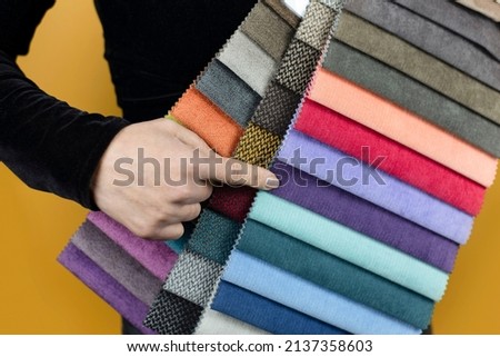 Catalog of fabric samples with different colors to choose fabric for upholstered furniture in the hands of woman. Woman points to purple fabric for her new sofa. Selective focus