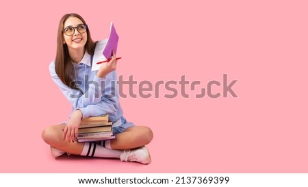 Thoughtful pretty blond school girl looking up with a smile sitting on the floor with crossed legs holding books and a diary with a pen, high school university concept. Royalty-Free Stock Photo #2137369399