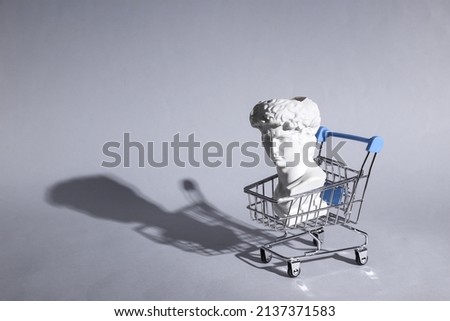 Antique bust of David with shopping trolley on gray background with shadow. Conceptual pop. Minimal shopping still life. Creative idea Royalty-Free Stock Photo #2137371583