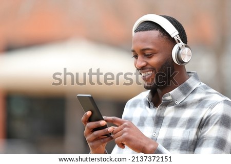 Happy man with black skin wearing headphones walking in the street listening to music checking smart phone