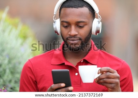 Front view portrait of a man with black skin wearing headphones watcking media on phone in a bar
