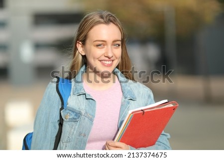 Happy student holding folders and a backpack walking looking at camera in a campus