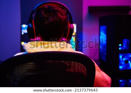 Back view of teenager boy play computer video game in dark room, use neon colored rgb mechanical keyboard, place for cybersport gaming
