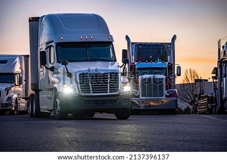 Different models of big rigs semi trucks tractors with car hauler and refrigerator and dry van semi trailers standing in row on the truck stop industrial parking lot for the rest at twilight time Royalty-Free Stock Photo #2137396137