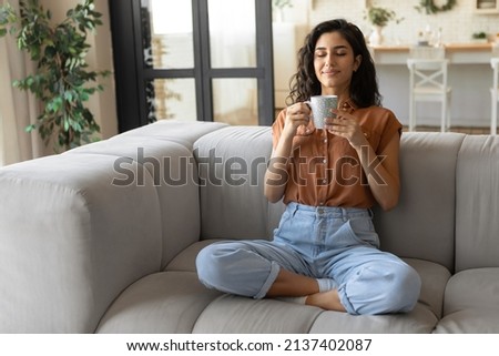 Peaceful young brunette woman sitting on couch with closed eyes, drinking hot coffee at home, copy space. Full length of millennial female enjoying lazy morning with warm drink in living room Royalty-Free Stock Photo #2137402087