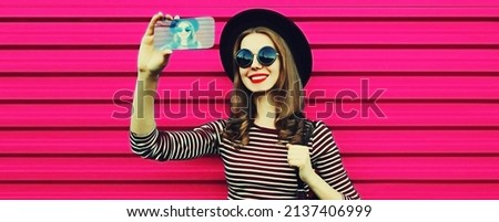 Portrait close up of happy smiling young woman taking selfie by smartphone wearing black round hat on pink background