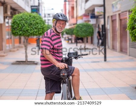 Senior smiling cyclist man wearing helmet cycling his electric bicycle in urban street stops looking back at camera. Active elderly grandfather enjoying a healthy lifestyle and freedom Royalty-Free Stock Photo #2137409529
