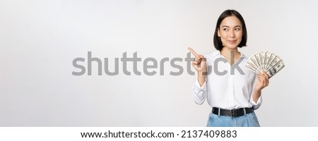 Smiling young modern asian woman, pointing at banner advertisement, holding cash money dollars, standing over white background Royalty-Free Stock Photo #2137409883