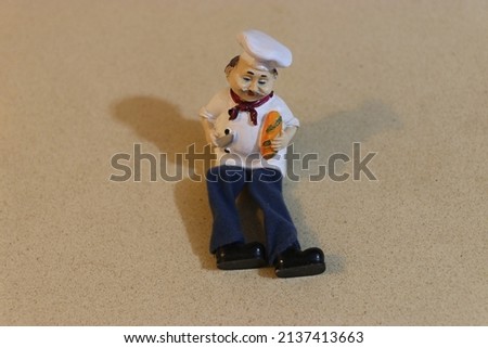 Chef porcelain doll on marble. Kitchen decoration with a game of silhouettes. A chef doll holding bread and a knife. A doll in a traditional chef's costume.Shelf doll