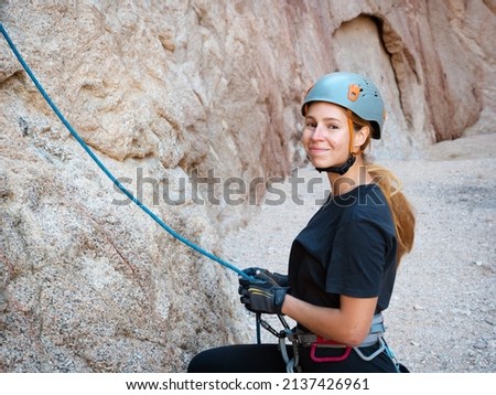 Rock climber in helmet stands in canyon, holds rope. Sportive girl smiling, looking at camera, keeping climbing equipment. Female mountaineer is preparing to climb mountain. Alpinist woman near cliff Royalty-Free Stock Photo #2137426961