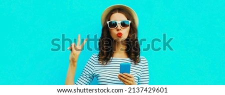 Portrait of happy young woman blowing her lips posing with smartphone wearing summer straw hat on blue background, blank copy space for advertising text